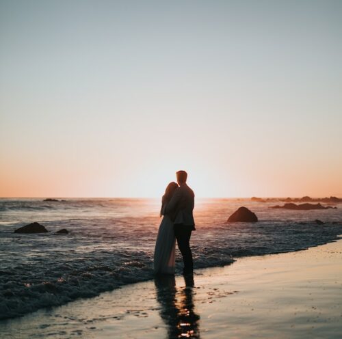 two people standing near the water on the beach while holding each other and watching the sunset